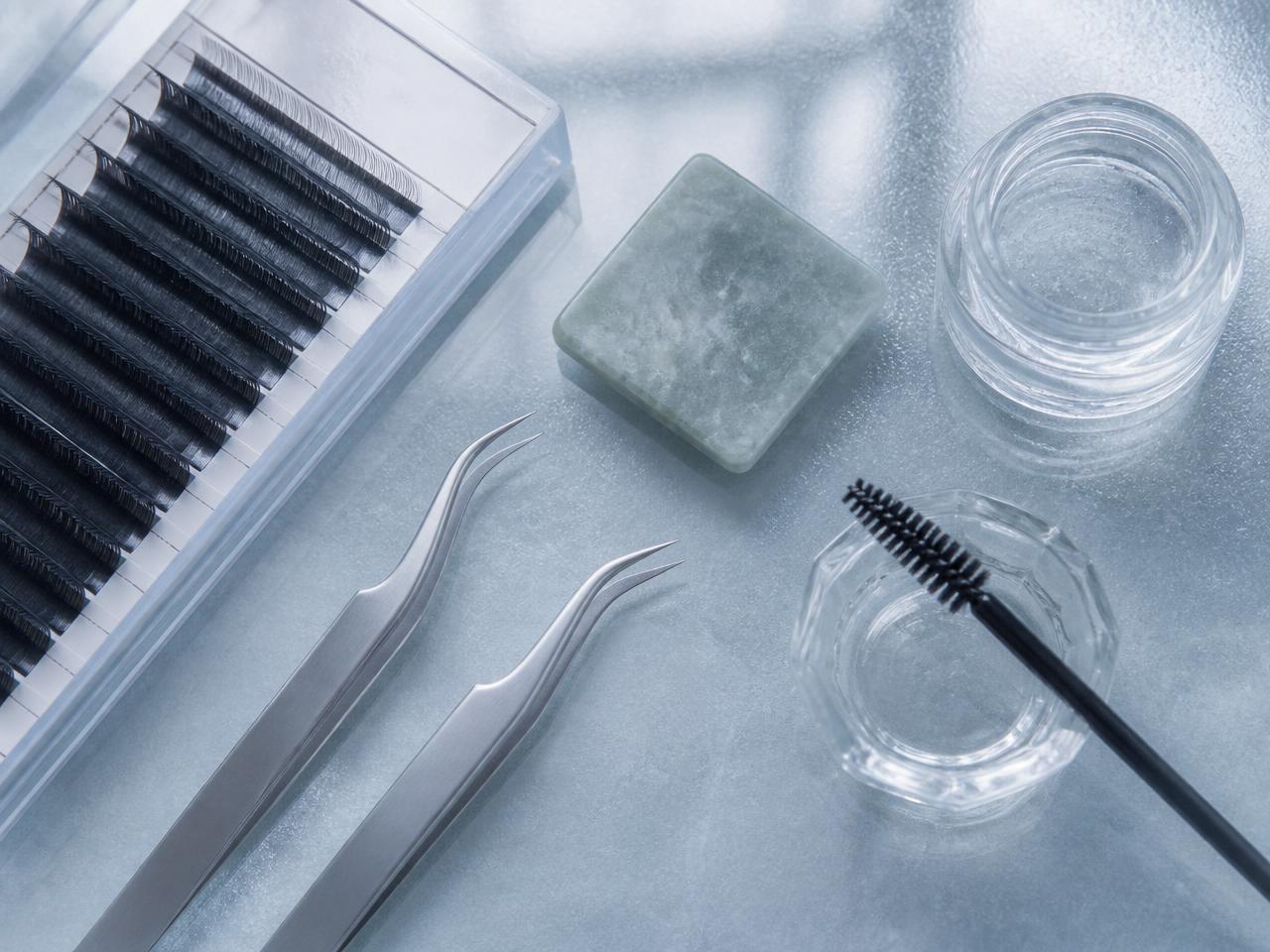 Fine curved tweezers, a tray of lash extensions and a brow lamination brush on a frosted glass surface